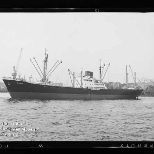 Schwarzweiß-Seitenansicht eines Frachtschiffs im Hafen vor Kränen und ruhiger Wasserfläche.