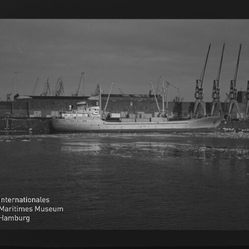 Frachtschiff am Kai vor Lagergebäuden und mehreren Hafenkränen, ruhige Wasseroberfläche mit Spiegelung.
