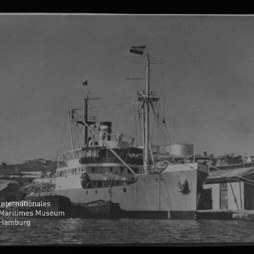 Großes Dampfschiff mit Schornstein und Masten am Kai, Bug mit Wappen und Gebäuden mit der Aufschrift 