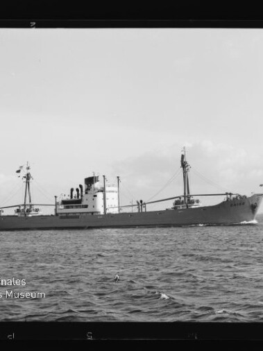 Frachtschiff in Seitenansicht auf ruhiger See mit Mast und Ladebaum, Museumslogo unten links.