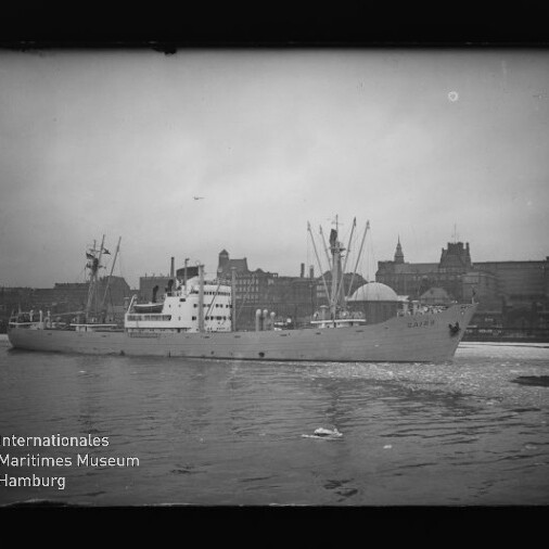 Frachtschiff mit Ladekränen fährt im Hafen vor Hamburger Stadtsilhouette; Museumslogo unten links.