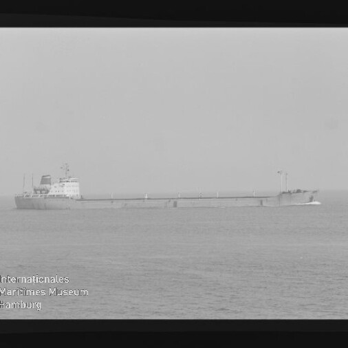 Schwarzweißfoto eines langen Frachtschiffs mit Aufbauten achtern auf ruhiger See; Filmrand und Museumslogo sichtbar.