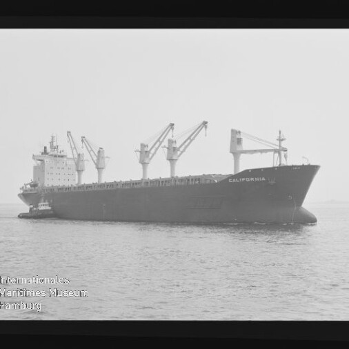 Massengutfrachter California mit mehreren Ladekränen auf ruhiger See, kleiner Schlepper am Heck.