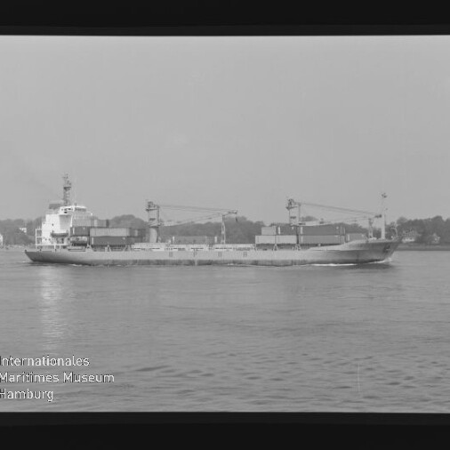 Seitliche Schwarzweißaufnahme eines Frachtschiffs mit Ladekränen und Containern vor dem Ufer, Filmrahmen mit AGFA PAN.