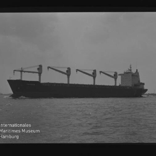 Schwarzweiß-Seitenansicht eines Frachtschiffs mit vier Ladekränen auf ruhiger See; Filmrand AGFA APX 100 und Museumslogo
