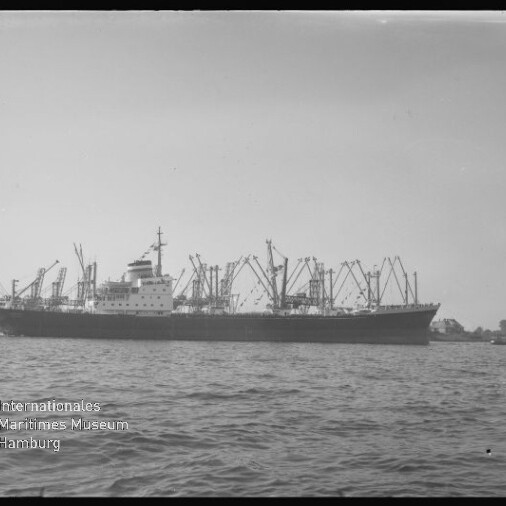 Großes Frachtschiff mit zahlreichen Ladekränen vor einem Hafen, ruhige See im Vordergrund und Museumssignet unten links.