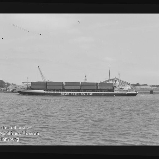 Frachtschiff mit aufgestapelten Containern und Schrift GEEST NORTH SEA LINE, vor Hafen mit Kränen, schwarz-weiß Aufnahme.