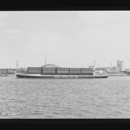 Schwarzweißfoto eines Frachtschiffs mit der Aufschrift GEEST NORTH SEA LINE, beladen mit Containern vor Hafenkränen.