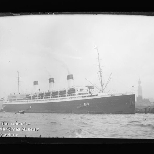 Großes historisches Passagierschiff mit drei Schornsteinen im Hafen, Wasser im Vordergrund und Glockenturm im Hintergrund.