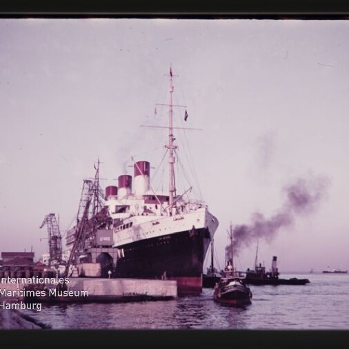Großer Ozeandampfer mit drei roten Schornsteinen am Kai neben Kränen, Schlepper mit Rauch und Museums‑Wasserzeichen