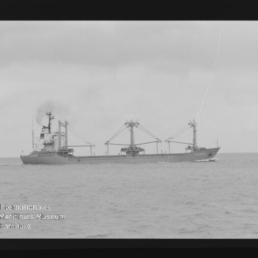 Schwarzweißfoto eines Frachtschiffs mit drei Ladekränen auf ruhiger See und Museumslogo links unten.