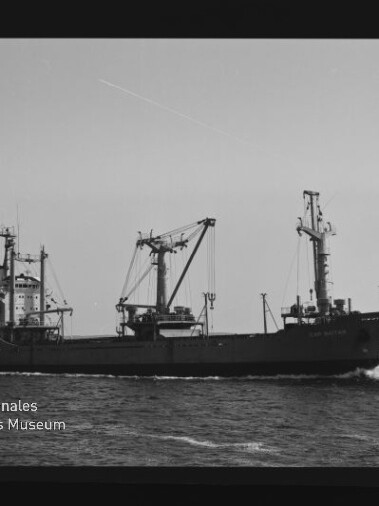Frachtschiff mit drei Ladekränen fährt auf ruhiger See, dunkler Filmrand und Museumslogo am linken unteren Bildrand sichtbar.