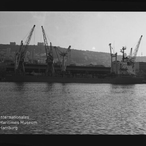 Schwarzweißfoto eines Frachtschiffs am Kai mit mehreren Kränen und Wasser im Vordergrund, unten links Museumslogo.