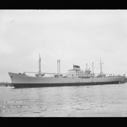 Schwarzweiß-Foto eines großen Frachtschiffs in Seitenansicht mit Mittschiffsaufbau, mehreren Masten und Schornstein, ruhig im Hafen liegend