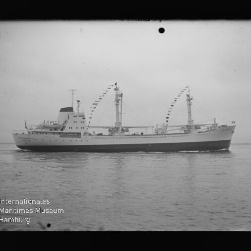 Frachtschiff mit zwei beflaggten Masten und sichtbarem Ladegeschirr, seitliche Ansicht auf ruhiger See.