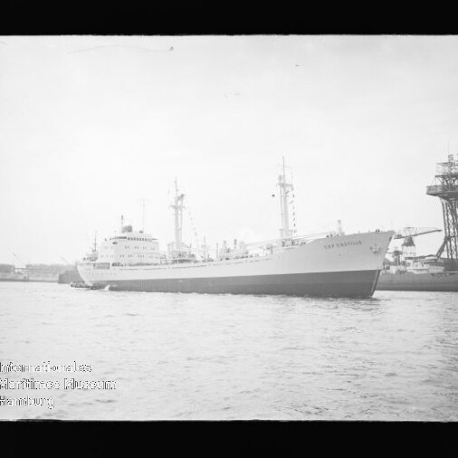 Schwarzweißaufnahme eines großen Frachtschiffs in Seitenansicht im Hafen, rechts ein hoher Werftkran und Industrieanlagen.
