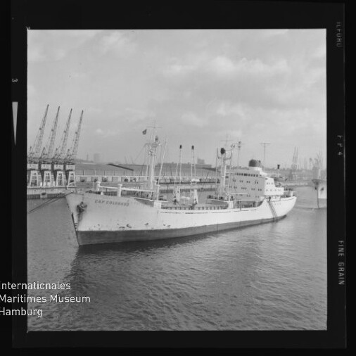 Frachtschiff Cap Colombie am Kai eines Industriehafens, mit Ladekränen und Hafenanlagen im Hintergrund.