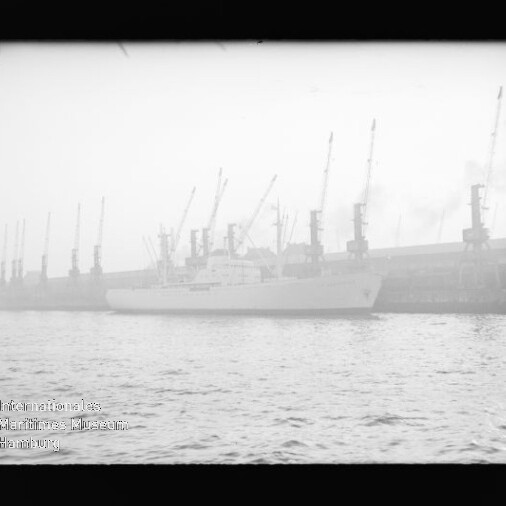 Weißes Schiff am Industriehafen mit einer Reihe hoher Kräne im Nebel, Wasser im Vordergrund und Museumsstempel links unten.