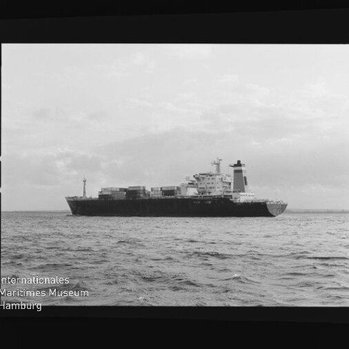 Frachtschiff mit gestapelten Containern fährt auf ruhiger See, Schwarzweißaufnahme mit Museumslogo unten links.