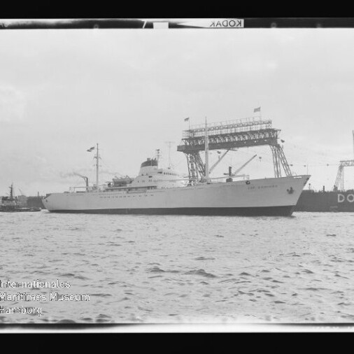 Großes weißes Passagierschiff vor Dockkran und Kaianlage, im Vordergrund Wasser und ein kleines Schlepperboot.