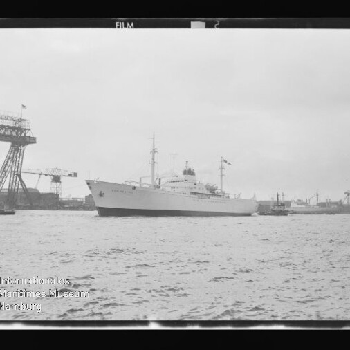Großes weißes Schiff mit Masten und Aufbauten liegt im Hafen, im Hintergrund Werftkräne und ruhiges Wasser.
