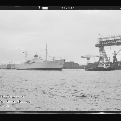 Weißes Schiff, begleitet von Schleppern, fährt an einem Werftgelände mit hohen Kranen und der Aufschrift 