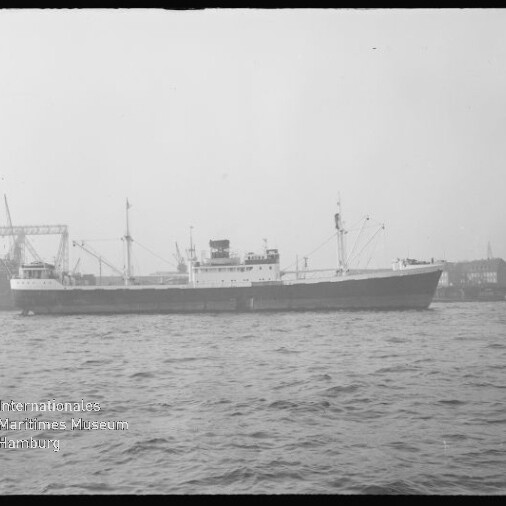 Seitliche Aufnahme eines Frachtschiffs im Hafen, Kräne und Werftgebäude im Hintergrund, Museumswasserzeichen links unten