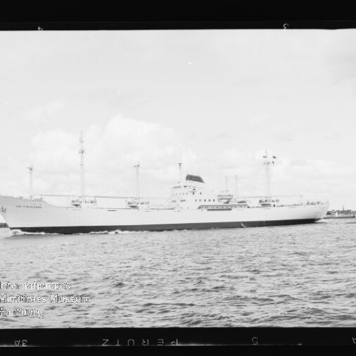 Weißes Frachtschiff mit mehreren Masten fährt auf ruhiger See, kleiner Schlepper am Heck.