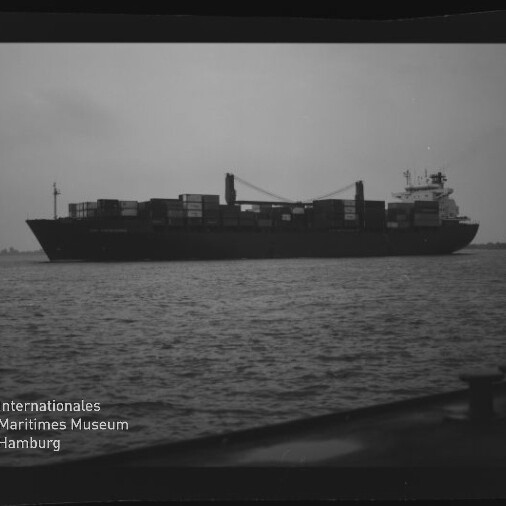 Schwarzweiß-Foto eines beladenen Containerschiffs mit zwei Ladekränen auf ruhiger See, Museumswasserzeichen unten links.