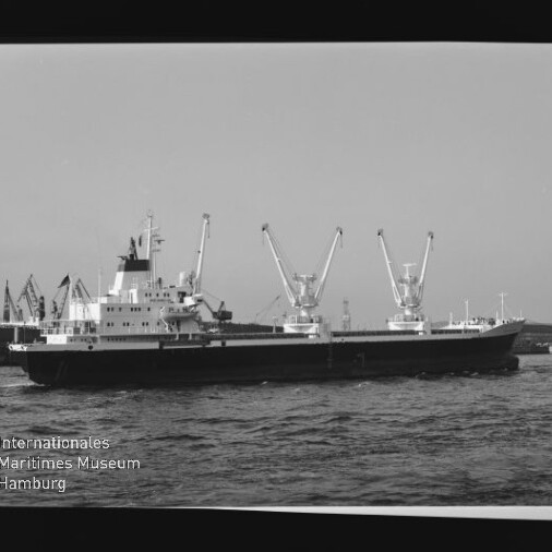 Frachtschiff mit drei Laderkränen auf dem Wasser vor Hafenanlagen; Wasserzeichen: Internationales Maritimes Museum Hamburg.