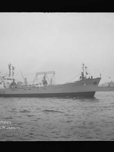 Schwarzweiß-Foto eines beflagten Frachtschiffs im Hafen vor Werftkränen, mit Wasser im Vordergrund und Museumslogo.