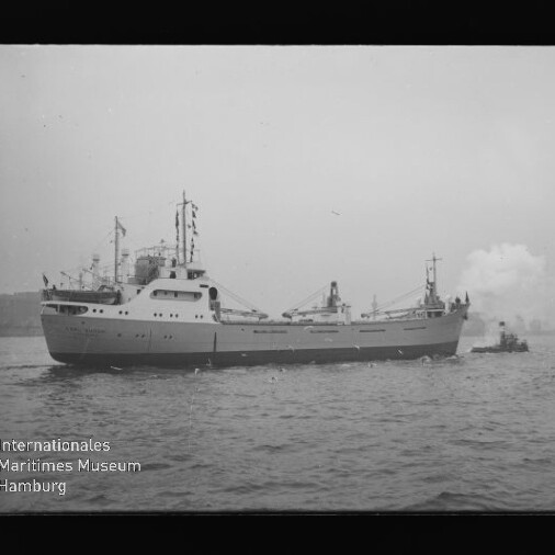 Frachtschiff mit vornliegenden Aufbauten und Flaggen, begleitet von kleinem Schlepper; sichtbares Museumslogo