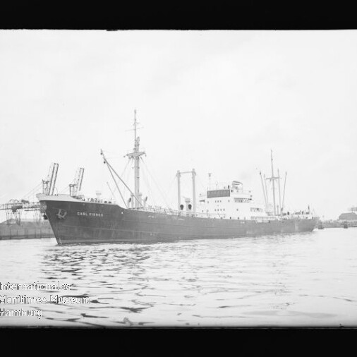Großes Frachtschiff vor Kaianlagen mit Hafenkränen, spiegelndes ruhiges Wasser.