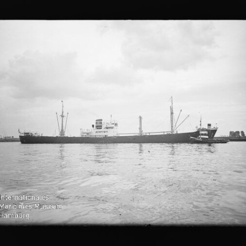 Langes Frachtschiff mit Schlepper im Hafen unter bewölktem Himmel; Wasseroberfläche und Museum-Logo links unten.