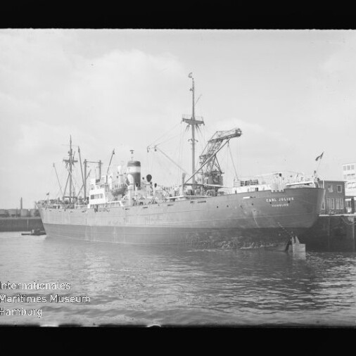 Großes Frachtschiff mit Deckskranen am Kai eines Industriehafens, ruhiges Wasser im Vordergrund.