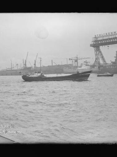 Kleines Frachtschiff auf dem Wasser vor einem Hafen mit zahlreichen Werftkränen und einem beschrifteten Portalkran.