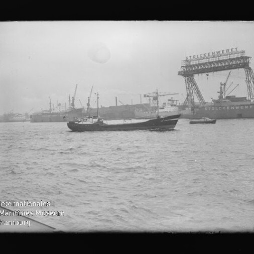 Kleines Frachtschiff auf dem Wasser vor einem Hafen mit zahlreichen Werftkränen und einem beschrifteten Portalkran.