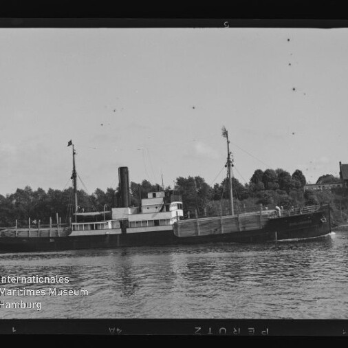 Schwarzweißfoto eines kleinen Dampfers mit Schornstein auf einem Fluss vor bewaldetem Ufer und Häusern.