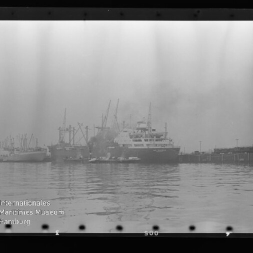 Mehrere angelegte Frachtschiffe und Kräne in nebligem Hafen, ruhige Wasserfläche; Museumssignet unten links.