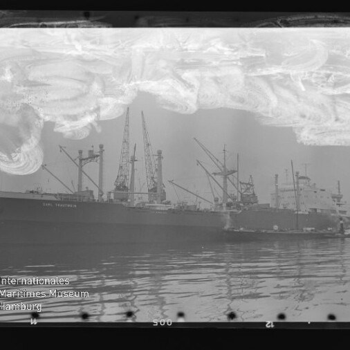 Großes Frachtschiff am Kai mit Ladekränen und Spiegelung im Wasser, Museumslogo unten links und Filmabnutzungen oben.