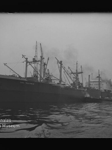 Großes Frachtschiff im Hafen neben einem weiteren Schiff und Hafenkränen, rauchende Schornsteine, Wasser im Vordergrund.