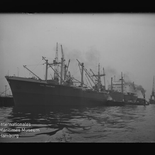 Großes Frachtschiff im Hafen neben einem weiteren Schiff und Hafenkränen, rauchende Schornsteine, Wasser im Vordergrund.