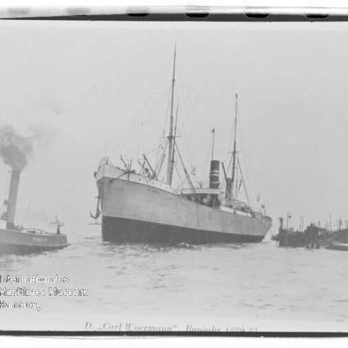 Schwarzweißaufnahme des Dampfschiffs „D. v. Carl Woermann“ mit hohem Schornstein und Masten, begleitet von einem Schlepper im Hafen.