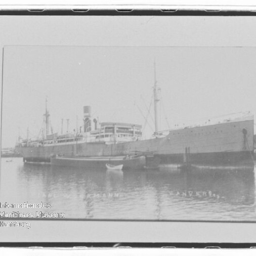 Schwarzweißaufnahme eines großen historischen Dampfschiffs am Kai, mit Spiegelung im Wasser und kleinem Ruderboot im Vordergrund.