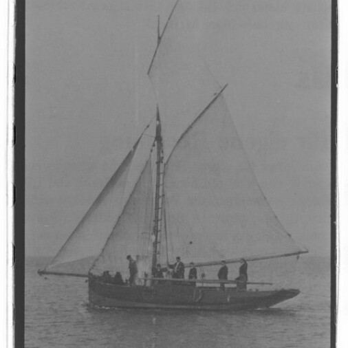 Segelboot mit zwei Segeln und mehreren Personen an Bord auf ruhiger See; Schwarzweißfoto mit Museumswasserzeichen
