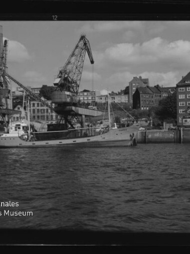 Frachtkahn am Kai unter zwei großen Hafenkränen, Häuser im Hintergrund und Wasser im Vordergrund; Museumssignet Internationales Maritimes Museum Hamburg links unten.