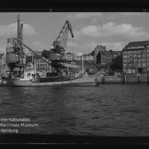 Frachtkahn am Kai unter zwei großen Hafenkränen, Häuser im Hintergrund und Wasser im Vordergrund; Museumssignet Internationales Maritimes Museum Hamburg links unten.