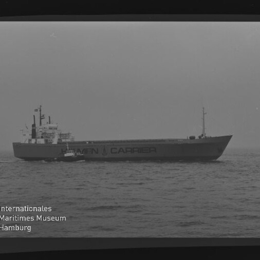 Großes Frachtschiff mit Schriftzug CARRIER auf ruhiger See, kleines Begleitboot links und Museumslogo unten links