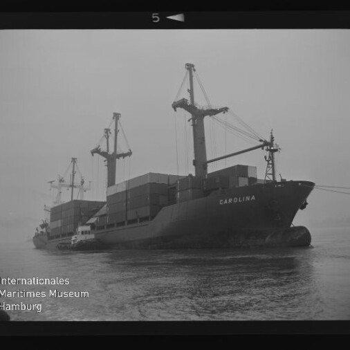 Frachtschiff CAROLINA mit Containern und Ladekränen, begleitet vom Schlepper, auf nebligem Wasser; Museumslogo unten links.