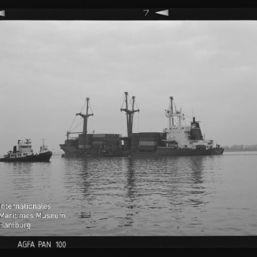 Frachtschiff mit Ladekränen und Containern, begleitet von einem Schlepper auf ruhigem Wasser; Schwarzweißaufnahme mit Museums-Wasserzeichen.